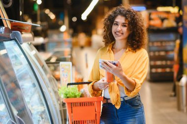 Gülümseyen kadın eşyaları kontrol ediyor ve parlak bir markette taze ürünlerle dolu bir alışveriş sepeti tutuyor.