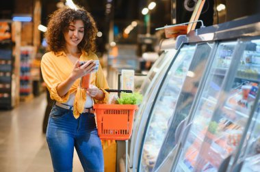 Genç bir kadın markette akıllı telefonuna bakarken gülümsüyor, elinde taze ürünlerle dolu bir alışveriş sepeti tutuyor.