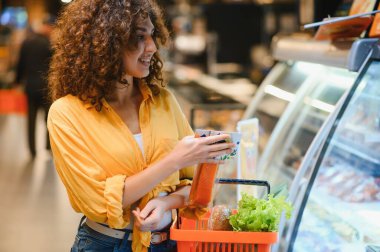 Gülümseyen, marketteki soğutuculu vitrinden paketli yiyecekleri seçen bir kadın.