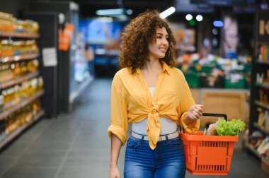 Parlak bir süpermarket koridorunda alışveriş sepetini taşırken gülümseyen genç bir kadın.