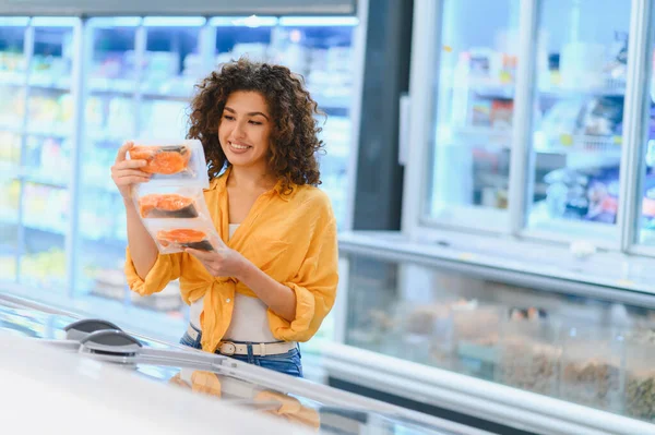 Gülümseyen kadın marketteki dondurucudan paketlenmiş somon alıyor, sağlıklı yemek seçeneklerini seçiyor.
