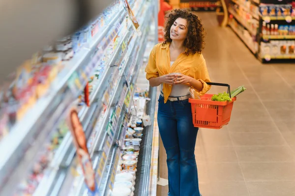 Kıvırcık saçlı, elinde alışveriş sepeti olan, rafları karıştıran ve marketten yiyecek seçen genç bir kadın.