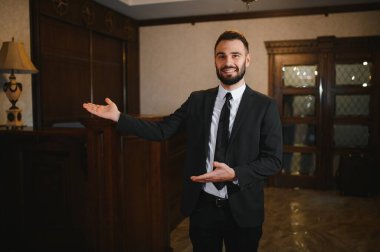 Gülümseyen sakallı adam resepsiyonist ya da kapıcı olarak çalışıyor. Lüks bir otel lobisinde konukseverlik hizmeti sunuyor.