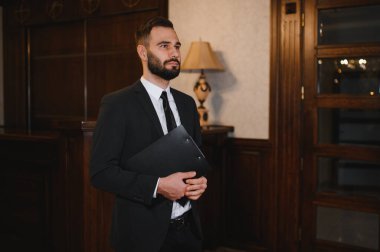 Siyah takım elbiseli bir adam elinde bir dosya, klasik bir otel lobisinde duruyor. Resepsiyonda profesyonel bir erkek var.