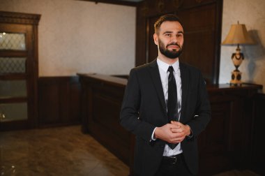 Otel lobisinde duran takım elbiseli ve kravatlı profesyonel bir adam. Mükemmel bir konukseverlik ve müşteri hizmetleri sunuyor.
