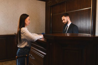 Otel resepsiyonunda duran bir misafir, bir erkek resepsiyoniste belgeleri veriyor, kontrol sürecini başlatıyor.