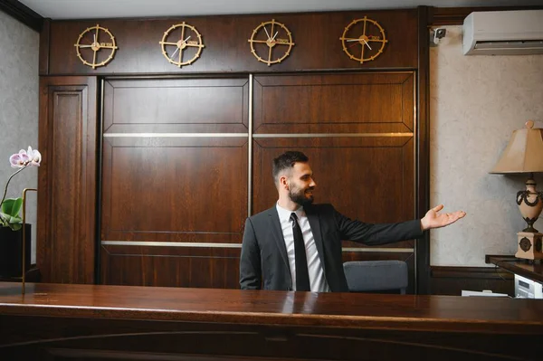 Gülümseyen otel kapıcısı ahşap bir resepsiyonda duruyor, misafirleri el hareketleriyle karşılıyor.