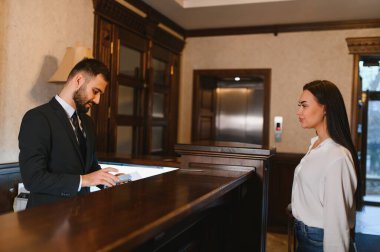 Resepsiyonist, resepsiyondaki bir bayana yardım ediyor. Otel lobisindeki prosedürleri tamamlıyor.