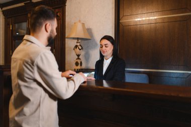 Adamın biri otel resepsiyonunda gülümseyen bir kadın resepsiyonistle görüşüyor.
