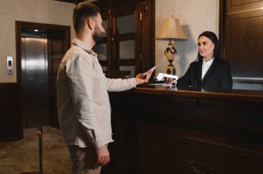 Otel resepsiyonunda ödemeyi akıllı telefondan yapan adam, dost canlısı bir bayan resepsiyonist tarafından tutulan NFC ödeme terminalini kullanıyor.