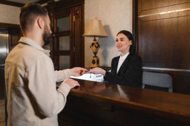 Otel resepsiyonisti resepsiyonistin resepsiyonda bir erkek misafirin anahtarlarını veriyor. Konukseverliğin altını çiziyor ve hizmet kaydını yaptırıyor.