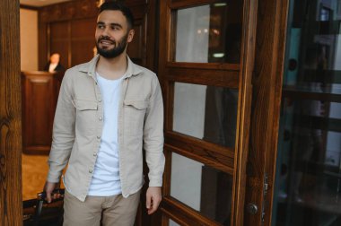 Otele sakallı bir adam giriyor, bavul getiriyor. Arkasında bir resepsiyonist olan ahşap bir kapının önünden başka tarafa bakıyor.