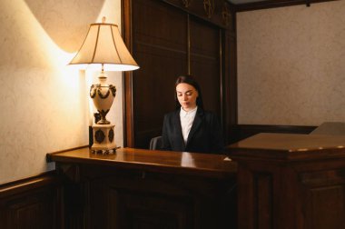 Bir otel lobisinde ahşap bir resepsiyonda çalışan takım elbiseli genç bir kadın profesyonel hizmet sunuyor.