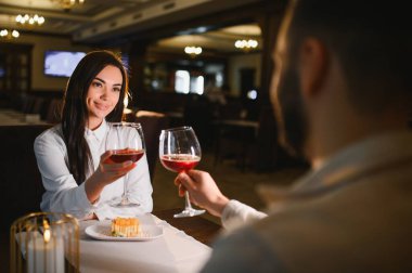 Genç bir kadın ve erkek, bir restoranda romantik bir akşam yemeğinde kırmızı şarap kadehleri kaldırarak özel bir olayı kutluyorlar.