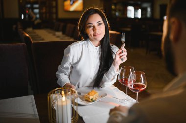 Genç bir kadın gülümsüyor, tatlı ve şarabı partneriyle paylaşıyor, yıldönümünü veya özel bir olayı karanlık bir restoranda kutluyor.