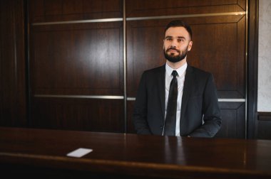 Otel lobisinde ahşap bir resepsiyon masasının arkasında duran takım elbiseli ve kravatlı bir adam profesyonel hizmet sunuyor.
