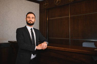 Otel resepsiyonunda duran takım elbiseli ve kravatlı profesyonel bir adam kameraya bakıyor ve gülümsüyor.