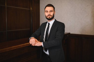 Sakallı ve takım elbiseli şık bir adam resepsiyon masasında duruyor. Otel lobisinde profesyonellik ve misafirperverlik yayıyor.