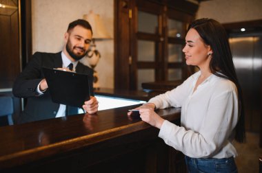 Gülümseyen otel resepsiyonisti genç bir bayana kayıt işlemlerini tamamlamasında yardımcı oluyor.