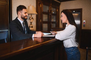 Otel resepsiyonisti, lüks bir otel lobisindeki resepsiyonda gülümseyen ve bir bayana yardım ediyor.