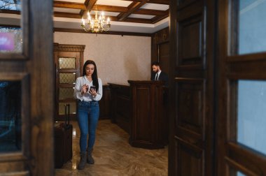 Otel resepsiyonuna gelen bir kadın, bavuluyla ayakta duruyor ve konaklaması için cep telefonuna yazı yazıyor.
