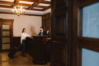 Bavulu olan kadın otel resepsiyonisti ile klasik bir lobide rahat, zarif bir resepsiyonda etkileşime geçiyor.