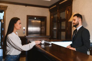 Misafir resepsiyonda kredi kartıyla ödeme yapıyor. Otel çalışanı giriş ve servise yardımcı oluyor.