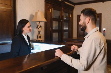 Dost canlısı otel resepsiyonisti, klasik bir otel lobisindeki resepsiyonda bir erkeğe yardım ediyor. Mükemmel bir hizmet ve konukseverlik sunuyor.