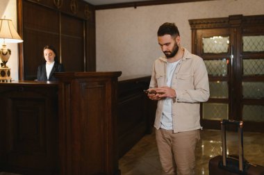 Bagajlı bir adam, otel resepsiyonunda akıllı telefonu kontrol ediyor, arka planda çalışan bir resepsiyonist. Seyahat ve konukseverlik kavramı