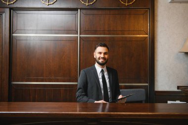 Takım elbiseli arkadaş canlısı resepsiyonist elinde bir dosya, resepsiyonda duruyor, misafirlere yardım etmeye hazır.