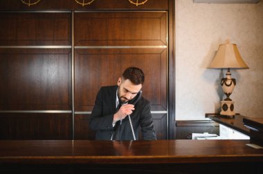 Erkek resepsiyonist otel lobisindeki bir müşteriye telefonla yardım ediyor. Mükemmel müşteri hizmetleri ve hoş geldin atmosferi sağlıyor.