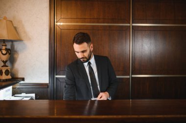 Profesyonel bir resepsiyonist ahşap tezgahın arkasında duruyor, bir otel veya iş lobisinde eşyalar düzenliyor.