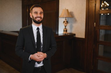 Klasik bir otel lobisinde gülümseyen erkek resepsiyonist. Mükemmel bir konukseverlik hizmeti sunarak dostça bir tutum sergiliyor.