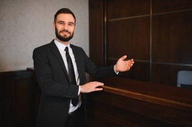 Takım elbiseli bir adam otel resepsiyonunda duruyor, gelen misafirleri selamlıyor. Profesyonel konukseverlik hizmeti sunuyor.