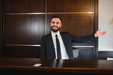 Dost canlısı otel resepsiyonisti ahşap bir tezgahın arkasında duruyor, gülümsüyor ve hoş geldin jesti yapıyor. Mükemmel bir konukseverlik ve hizmet sunuyor.