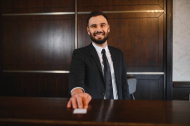 Erkek otel resepsiyonisti gülümsüyor, resepsiyonda anahtar kartı sunuyor, otel lobisinde dostça hizmet veriyor.
