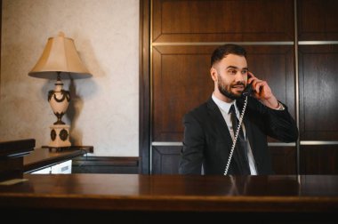 Profesyonel otel resepsiyonisti. Müşteri hizmetleri sunuyor. Zarif resepsiyonun arkasındaki sabit telefon görüşmesine cevap veriyor.