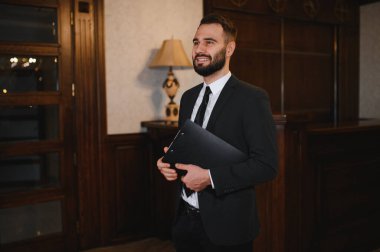 Takım elbiseli gülümseyen bir adam elinde bir pano bir otel lobisindeki resepsiyon masasında duruyor, konukseverliği resmediyor.