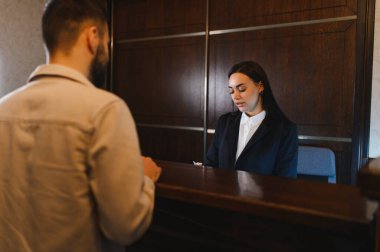 Genç bir adam otel lobisindeki resepsiyonistle görüşüyor. Konukseverlik hizmeti veriyor.