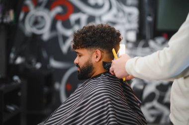 Saç ve sakallı genç bir adama şık bir saç kesimi yapmak için kırpıcı ve tarak kullanan berber stilisti.