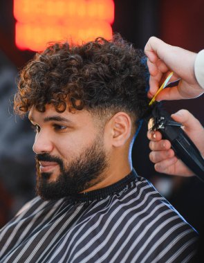 Kıvırcık saçlı ve sakallı genç adam elektrikli makas ve tarak kullanarak berberden saç tıraşı oluyor. Profesyonel tımar hizmeti.