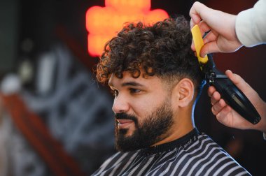 Kıvırcık saçlı ve sakallı bir adam ileriye bakarken berber saç kesme makinesi ve saç şekillendirme için tarak kullanıyor.