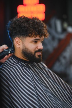 Kıvırcık saçlı genç adam bir berberden saç kesimi alıyor, budama ve şekillendirmeye odaklanıyor.