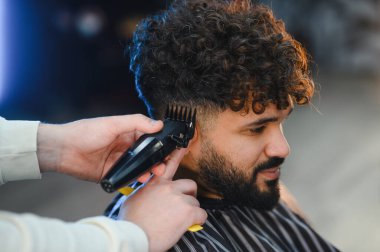 Profesyonel berber, modern bir kuaförde saç makasıyla müşterinin kıvırcık saçlarını buduyor ve yeni bir görünüm yaratıyor.