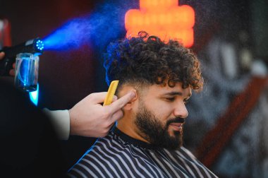 Berber, kıvırcık saçlı ve sakallı bir genci saç kesimi için hazırlıyor, su püskürtüyor ve berber dükkanında tarakla şekil veriyor.