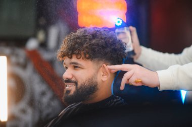 Kıvırcık saçlı, gülümseyen adam yeni saç kesimi alıyor ve modern bir berber dükkanında stil yapıyor. Tedavinin tadını çıkarıyor.