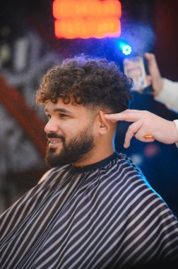 Mutlu müşteri gülümserken kuaför sprey sıkar ve kıvırcık saçlarını profesyonel bir berber dükkanında şekillendirir.