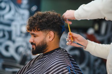 Erkek müşteri yeni saç stilini beğeniyor, kıvırcık saçlarını makas ve tarak kullanan bir stilist kestiriyor.