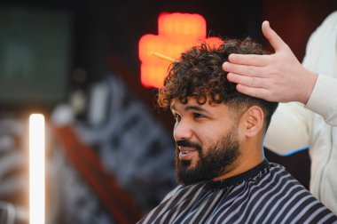 Kıvırcık saçlı ve sakallı genç adam kuaförde berberden profesyonel bir saç kesimi alırken gülümsüyor.
