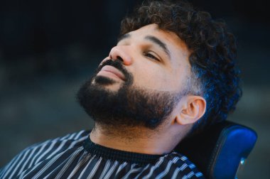 Kıvırcık saçlı ve sakallı genç adam berber dükkanında sakalını keserken bir anlık rahatlama ve özen gösteriyor.